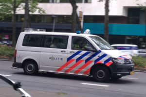 Ein Polizeiwagen fährt auf einer Straße neben einem Fahrrad, mit Menschen im Wagen und einem Gebäude, Bäumen und Pflanzen im Hintergrund.