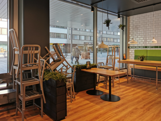 Ein Restaurant in Berlin mit Holztischen und -stühlen vor einem großen Fenster, umgeben von Pflanzen und Lichtern, mit Fußgängern und Gebäuden sichtbar draußen.