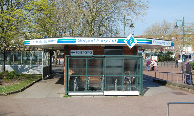 Haltestelle mit einem "Gosport Ferry Ltd"-Schild, Metallzaun, Menschen, Fahrrädern, Bäumen, Straßenlaternen, Schildern, einem Gebäude und einem bewölkten Himmel.