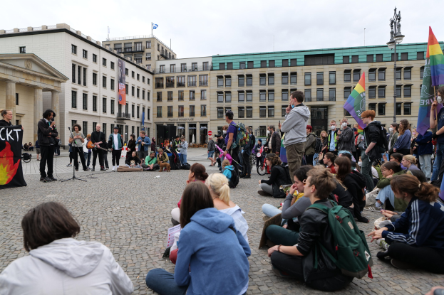 Eine Gruppe von Menschen, die auf dem Boden vor einer Menge sitzen, die Fahnen und Transparente hält, während einer Anti-Schwulen-Demo in Berlin. Im Hintergrund sind eine Statue, Gebäude und ein bewölkter Himmel zu sehen.