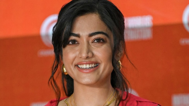 Eine Frau in einem roten Kleid mit einem eleganten Dutt und dezentem Make-up steht lächelnd da, trägt schwarze High Heels und hat ein hellgelbes Banner mit der Aufschrift "Sonam Kapoors beste Beauty-Looks vom Cannes Film Festival" im Hintergrund.