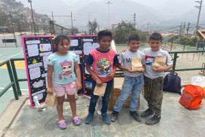 Eine Gruppe von Kindern steht auf einem Basketballfeld, jedes hält einen Papiertüte, mit Tüten und Gegenständen rechts und einer Texttafel links, mit Zaun, Pfosten, Straßenlaternen, Strommasten mit Drähten, Bäumen, Gebäuden, Hügeln und dem Himmel im Hintergrund.