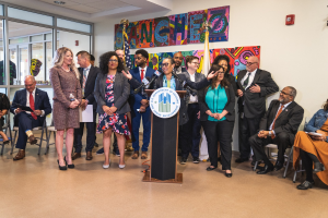 Gruppe von Menschen vor einem Podium mit Mikrofon und einer Tafel mit Text, mit sitzenden und stehenden Zuschauern, Fahnen, Feuerlöscher und Deckenleuchten im Hintergrund.