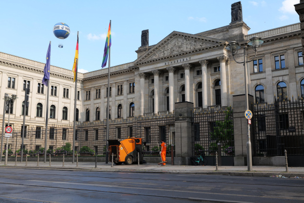Großes modernes Gebäude mit vielen Fenstern, als Bundestag in Berlin, Deutschland identifiziert, umgeben von Straßeninfrastruktur und Fußgängern, mit Flaggen und Bäumen im Vordergrund und einem bewölkten Himmel im Hintergrund.