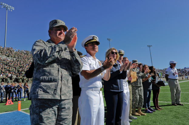 Eine Gruppe von Menschen auf einem Fußballfeld stehend und klatschend, mit Zuschauern auf den Tribünen, Flutlichtmasten und einem klaren blauen Himmel im Hintergrund.
