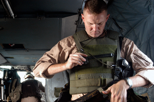 Ein Mann in militärischer Uniform steht im hinteren Teil eines Flugzeugs, hält eine Waffe in einer Hand und eine Tasche in der anderen und trägt eine kugelsichere Weste.