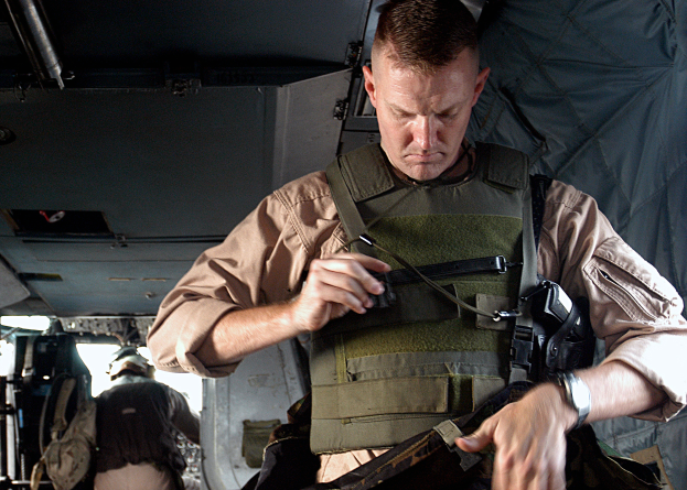 Ein Mann in militärischer Uniform steht im hinteren Teil eines Flugzeugs, hält eine Waffe in einer Hand und eine Tasche in der anderen und trägt eine kugelsichere Weste.