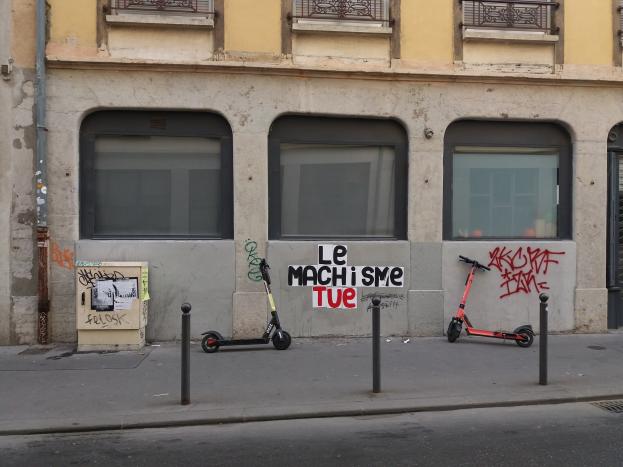 Zwei Roller auf einer Straße neben einem Gebäude mit Fenstern, Gelöndern und einem Rohr geparkt, mit Masten und einer Texttafel in der Nähe.