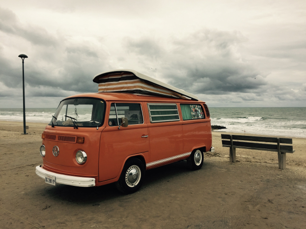 Oranger VW-Bus auf einem sandigen Strand nahe dem Ozean geparkt, mit einer h├Âlzernen Bank und einem Laternenpfahl rechts daneben, unter einem bew├Âlktem Himmel mit dem Wasser im Hintergrund.