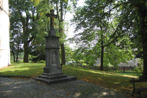 Ein Denkmal steht in einem Park mit einer Bank auf seiner rechten Seite, umgeben von Gras, Bäumen und einem Gebäude im Hintergrund unter einem klaren Himmel.