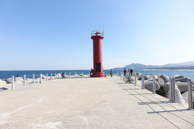 Roter Leuchtturm auf einem Pier am Meer mit Menschen, Fahrrädern, Geländern und Felsen in der Nähe, Hügel im Hintergrund unter einem klaren blauen Himmel.