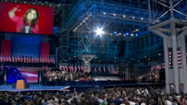 Eine große Menschenmenge sitzt vor einer Bühne mit einem Rednerpult, Mikrofon, Fahnen, einem Bildschirm mit dem Bild einer Frau und Beleuchtungsausrüstung und wartet auf eine Präsidentschaftsdebatte.