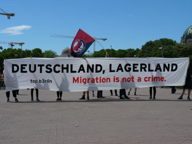 Eine Gruppe von Menschen steht draußen und hält ein Banner mit der Aufschrift "Deutschland, Lagerland Migration ist kein Verbrechen" und eine Flagge, mit Straßenlaternen, Bäumen, Kränen, einem Gebäude und einem bewölkten Himmel im Hintergrund.