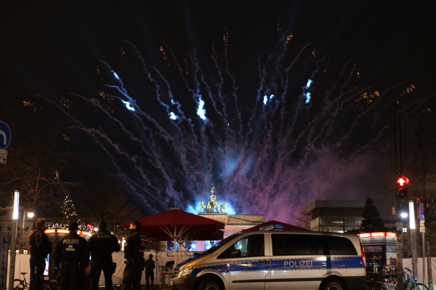 Ein Polizeiwagen steht vor einer Menschenmenge, umgeben von Fahrrädern, Zelten, Laternenmasten, Schildern, Verkehrszeichen, Bäumen, Gebäuden und einer Statue, unter einem von Feuerwerk erhellten Himmel.