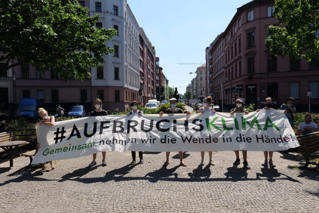 Eine Gruppe maskierter Individuals mit einer Banner tragend 'Aufbruchsklima' protestiert gegen den Klimawandel in Berlin, Deutschland, mit Gebäuden, Bänken, Tischen, Pflanzen, Bäumen, Fahrzeugen, Laternenpfählen und Schildern im Hintergrund.