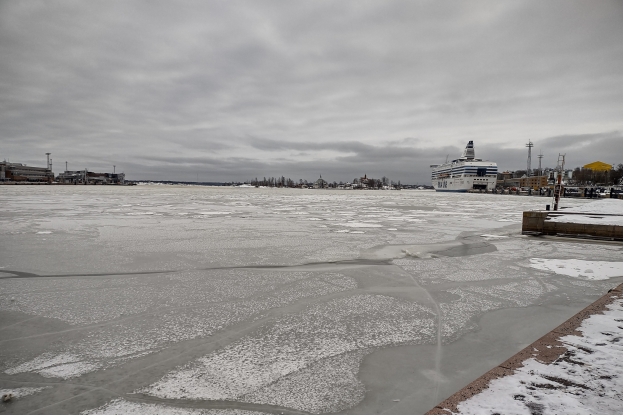 Großes Kreuzfahrtschiff liegt nahe einer verschneiten Wasserfront mit Gebäuden, Bäumen und Masten im Hintergrund unter einem bewölkten Himmel.