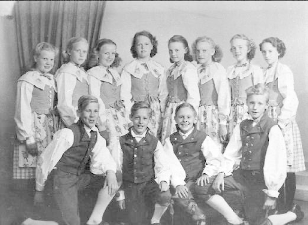 Schwarzes und weißes Foto einer Gruppe von Kindern, die vor einer Wand mit einem Vorhang posieren, mit dem Text "1930er-Kinder-Schule" unten.