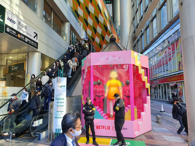 Eine Gruppe von Menschen steht vor einem Gebäude mit Glaswänden und Texttafeln, einige tragen Masken, während eine Rolltreppe mit Menschen zu sehen ist; ein pinker Kasten in der Mitte zeigt "Netflix's new Netflix booth in Tokyo" mit Schaufensterpuppen darin.