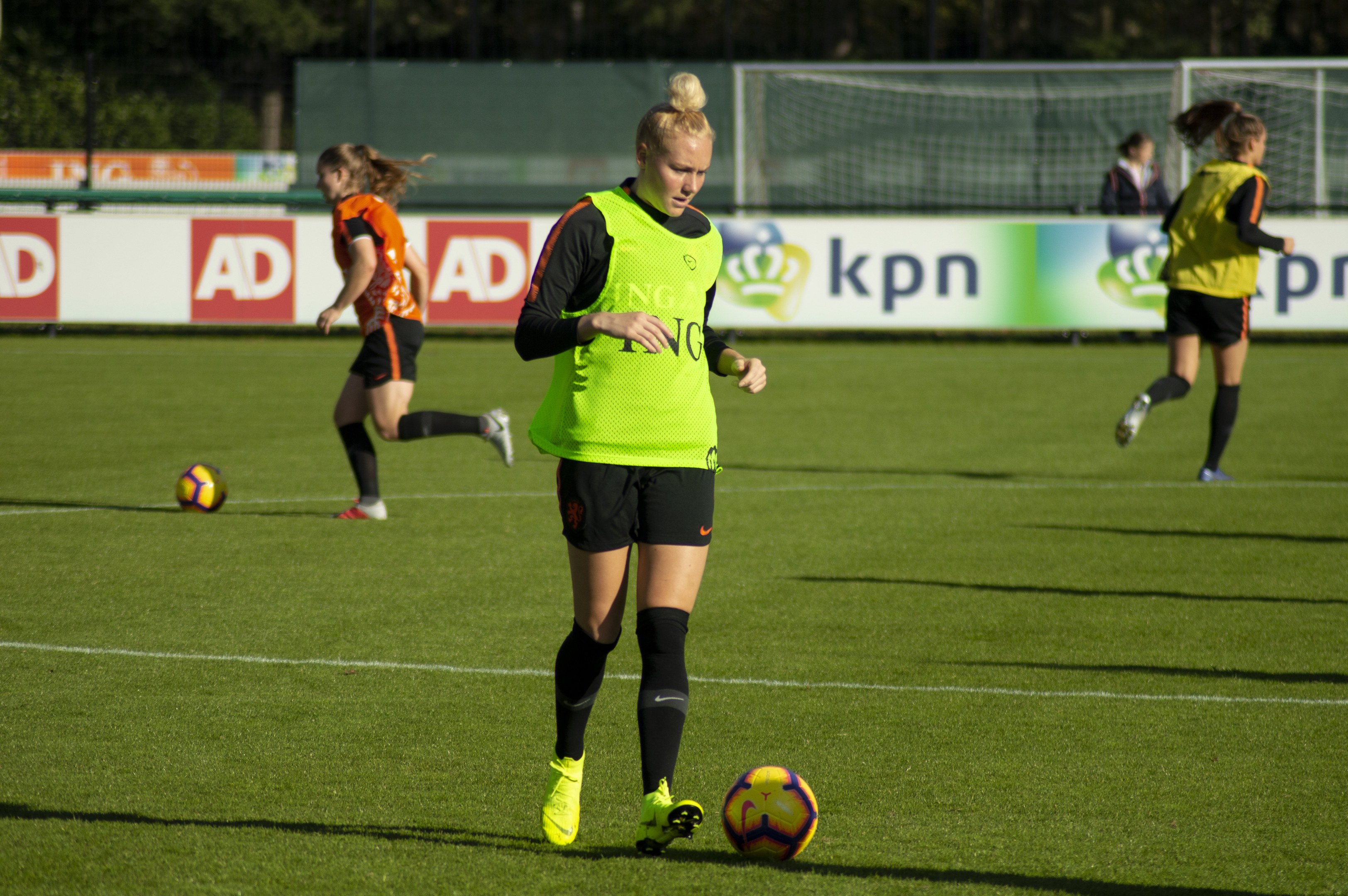 Gruppe von Frauen in Fußballtrikots beim Spiel auf einem Feld mit Bäumen im Hintergrund, Bannern dahinter und einem Netz im Vordergrund.