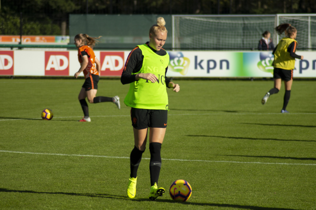 Gruppe von Frauen in Fußballtrikots beim Spiel auf einem Feld mit Bäumen im Hintergrund, Bannern dahinter und einem Netz im Vordergrund.