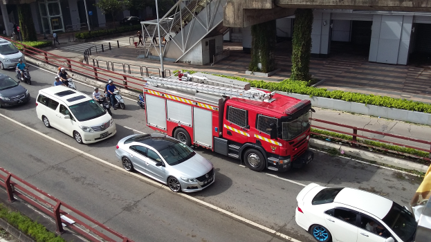 Ein Feuerwehrauto fährt auf einer Straße neben einem Auto, mit Motorradfahrern und anderen Fahrzeugen im Hintergrund.
