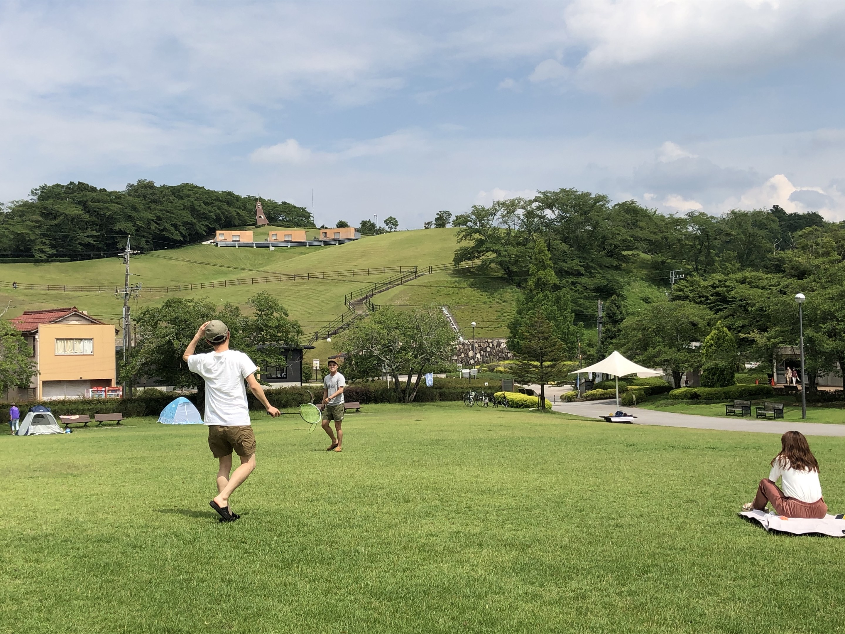 Menschen spielen Badminton in einem Park mit Zelten und Gebäuden im Hintergrund.