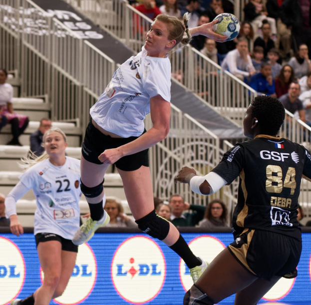 Eine Gruppe von Frauen beim Handballspielen auf einem Platz mit einem Ball in der Luft, Zuschauer auf Treppen und Geländern im Hintergrund und ein Schild mit der Aufschrift "Handball-Weltmeisterschaft 2019 - Tag 2".