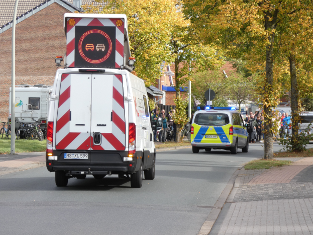 Ein Krankenwagen fährt eine von Bäumen gesäumte Straße mit Gebäuden entlang, während Menschen auf dem Gehweg neben Fahrrädern, Pfählen und Gras stehen.