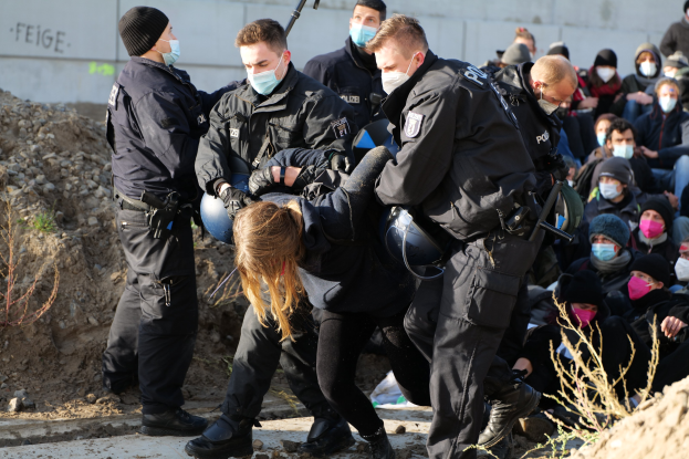 Eine Gruppe maskierter Personen in Mützen, einige bewaffnet, umgibt eine Frau, die von der Polizei weggeführt wird, mit Pflanzen, Steinen und einer Wand mit Protesttext im Hintergrund.