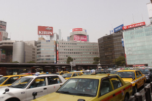 Eine Stadtansicht mit Fahrzeugen auf der Straße, Gebäuden, Bäumen, Pfählen mit Schildern und Text auf den Schildern, einem Zug im Hintergrund und einem Geländer unten rechts.