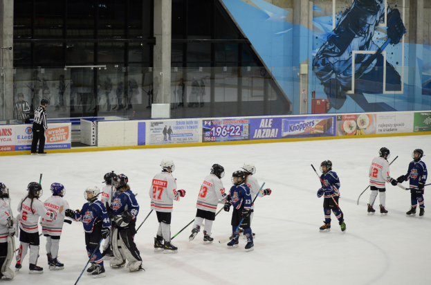Eine Gruppe junger Menschen, die Eisockey auf einer Indoor-Eisfläche spielen, tragen Helme und halten Stöcke, mit einer Person links daneben und Bannern, Säulen und Glaswänden im Hintergrund.