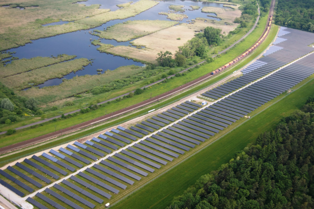 Luftaufnahme einer Solaränlage mit Panelen, umgeben von Bäumen, Gras und Wasser, mit einem Zug auf einer nächsten Eisenbahnschiene.