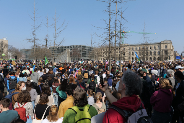 Eine große Menge Menschen mit Schildern und Taschen vor einem Gebäude mit Fenstern, Bäumen und einem klaren Himmel im Hintergrund, was auf eine Klimaprotestveranstaltung hinweist.