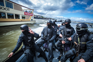 Eine Gruppe von Polizisten in schwarzen Uniformen und Helmen, einige bewaffnet, fährt auf einem Boot auf dem Wasser mit einer Brücke, Bäumen, Gebäuden und einem klaren blauen Himmel im Hintergrund.
