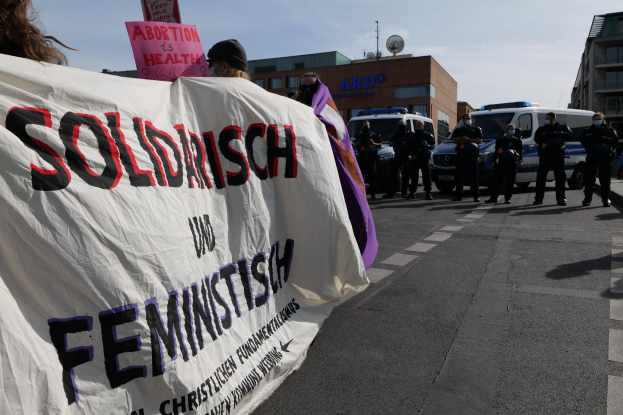 Gruppe von Menschen marschiert mit einem "Solidarität und Feminismus"-Plakat die Straße entlang, mit parkenden Fahrzeugen und Gebäuden im Hintergrund bei klarem blauem Himmel.