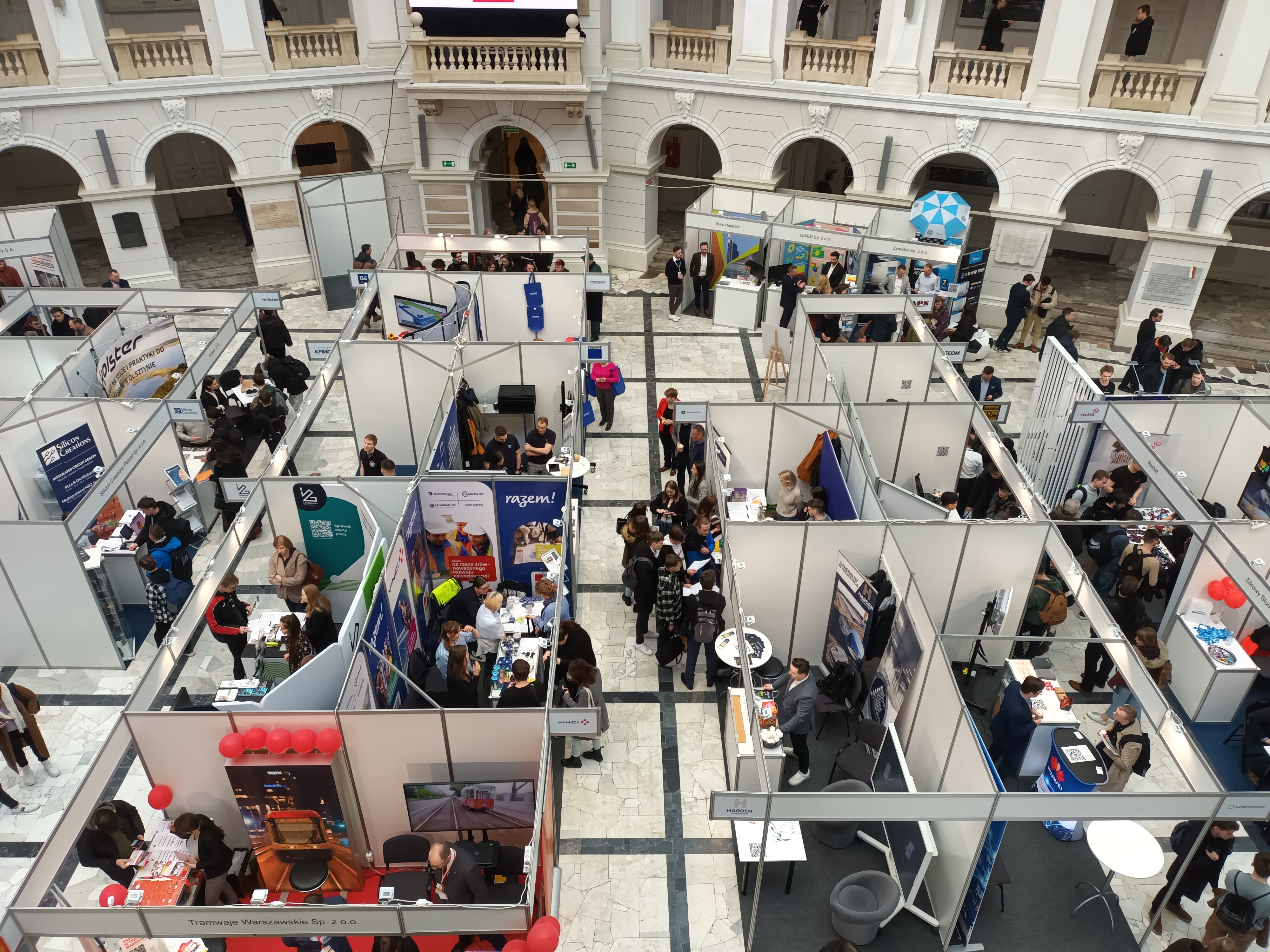 Eine große Gruppe von Menschen versammelte sich in einem Raum voller Stände, Banner, Tische, Stühle, Luftballons und einem Gebäude mit Bögen, Säulen, Geländern und einem Bildschirm im Hintergrund.