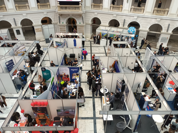 Eine große Gruppe von Menschen versammelte sich in einem Raum voller Stände, Banner, Tische, Stühle, Luftballons und einem Gebäude mit Bögen, Säulen, Geländern und einem Bildschirm im Hintergrund.