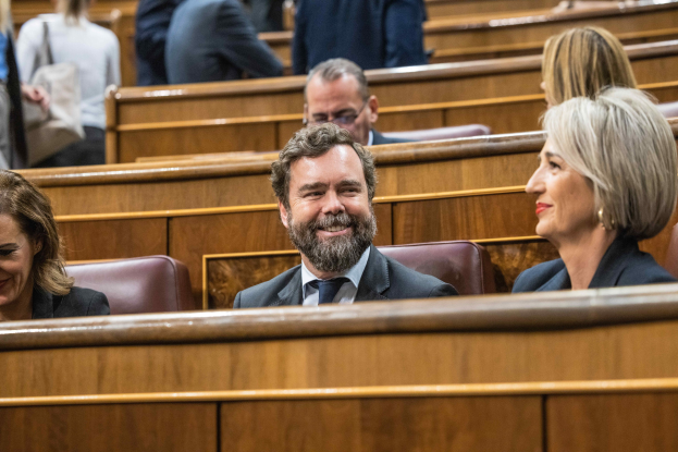 Gruppe von Menschen, die in einem Gerichtssaal sitzen, mit einem Mann in Anzug und Krawatte und zwei leger gekleideten Frauen, die nebeneinander sitzen, sowie sichtbaren Bänken im Hintergrund.