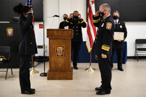 Gruppe uniformierter Personen auf einem Podium bei einer Vereidigung eines neuen Feuerwehrchefs, der eine Urkunde hält, mit Flaggen und Sitzplätzen im Hintergrund.