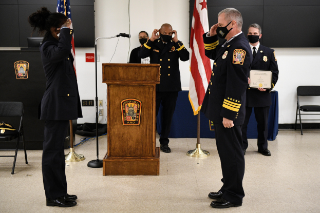 Gruppe uniformierter Personen auf einem Podium bei einer Vereidigung eines neuen Feuerwehrchefs, der eine Urkunde hält, mit Flaggen und Sitzplätzen im Hintergrund.