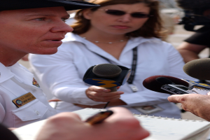 Eine Frau in einem weißen Kleid und schwarzem Hut spricht mit Reportern, während rechts ein Mann in einem schwarzen Hemd eine Kamera hält, ein Buch vor ihnen und mehrere Menschen im Hintergrund.