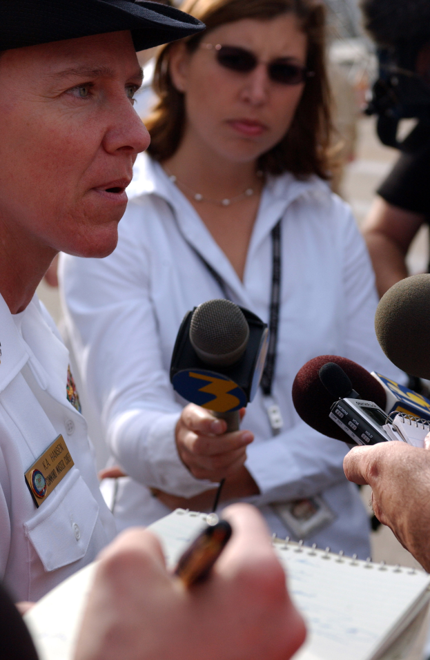 Eine Frau in einem weißen Kleid und schwarzem Hut spricht mit Reportern, während rechts ein Mann in einem schwarzen Hemd eine Kamera hält, ein Buch vor ihnen und mehrere Menschen im Hintergrund.