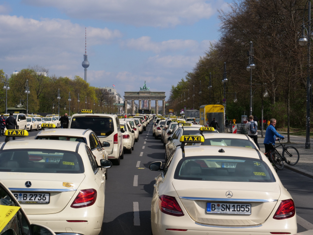 Eine lange Reihe von Taxis, die entlang einer belebten Straße in Berlin, Deutschland, geparkt sind, mit Fahrradfahrern und Fußgängern auf dem Bürgersteig, flankiert von Bäumen und Laternen, und Gebäuden, einem Bogen und einem Turm im Hintergrund unter einem bewölkten Himmel.