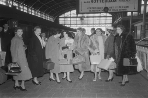 Schwarzes und weißes Bild einer Gruppe von Frauen, die an einem Bahnhof stehen, jede hält eine Tasche, mit einem Zug auf der linken Seite und Geländern und einer Tafel auf der rechten Seite.