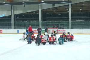 Eine Gruppe von Kindern in Helmen und mit Hockey-Schlägern sitzt auf einem Eisstadion, mit einer Wand aus Glas und Säulen, Deckenlampen und Texttafeln im Hintergrund.
