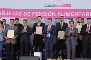 Eine Gruppe von Menschen auf einer Bühne, die klatschen und Zertifikate halten, mit Stühlen in einem Halbkreis und einem Banner mit der Aufschrift "Capital Social for Ti" im Hintergrund sowie Blumensträuße und Redner am Boden.