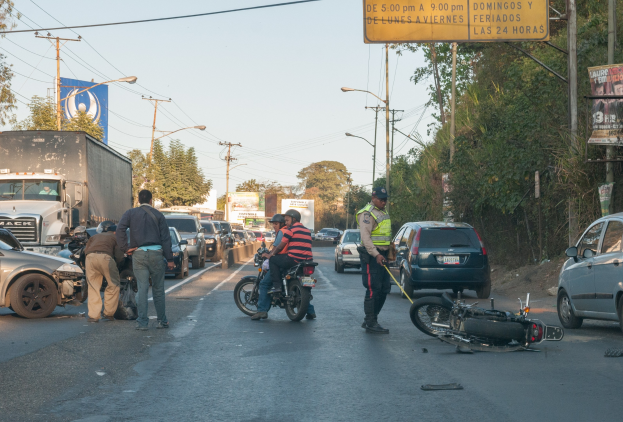 Eine Gruppe von Menschen steht um ein verunglücktes Motorrad am Straßenrand mit mehreren Fahrzeugen, Bäumen, Masten, Lichtern, Schildern und dem Himmel im Hintergrund.