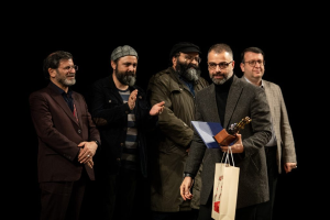 Eine Gruppe von Männern steht auf einer Bühne zusammen, einer hält eine Tasche und eine Trophäe, mit einem dunklen Hintergrund, der die Eröffnung der Berliner Filmfestspiele suggeriert.