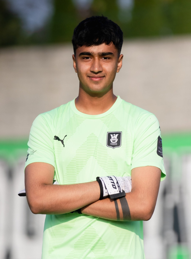 Ein junger Mann in einem grünen T-Shirt steht auf einem Fußballfeld und verschränkt die Arme, während er lächelt, mit einem unscharfen Hintergrund.