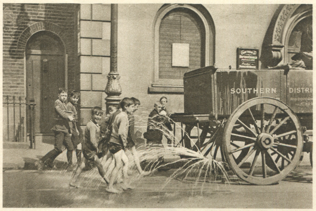 Schwarzes und weißes Bild von Kindern, die mit einem Feuerhydranten vor einem Gebäude mit Fenstern, einer Tür, einem Mast und einem Zaun spielen, mit einem Wagen im Hintergrund.